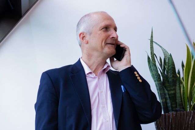 A man in a blue suit and pink shirt holds a smartphone to his ear and smiles. A succulent can be seen in the background on the right.