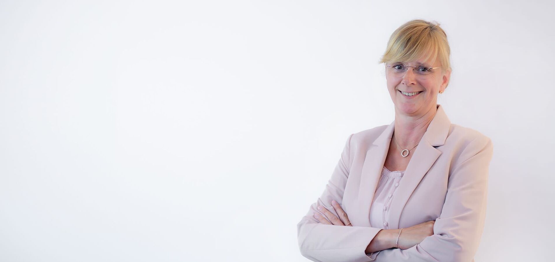 A portrait photo of a middle-aged lady in front of a light gray wall. She wears a blouse and a blazer in rosé and crosses her arms. She has her blond hair tied up, wears glasses and smiles at the camera.