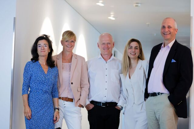 Auf diesem Teamfoto im Büro lächeln von links zwei Frauen, ein Mann, eine weitere Frau und wieder ein Mann. Sie haben unterschiedliche Haltungen und wirken sehr kompetent.