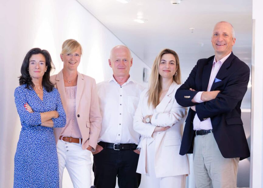 Auf diesem Teamfoto im Büro lächeln von links zwei Frauen, ein Mann, eine weitere Frau und wieder ein Mann. Sie haben unterschiedliche Haltungen und wirken sehr kompetent.