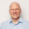 Portrait photo of DP-Dock shareholder: Dr. Paul Voigt in front of a white wall. He is wearing a light blue shirt and smiling.