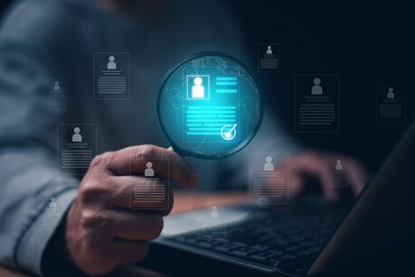 Hand hält eine Lupe über ein digitales Personenprofil mit Häkchen, umgeben von Profil-Icons vor einem Laptop – Symbol für die Suche nach personenbezogenen Daten
