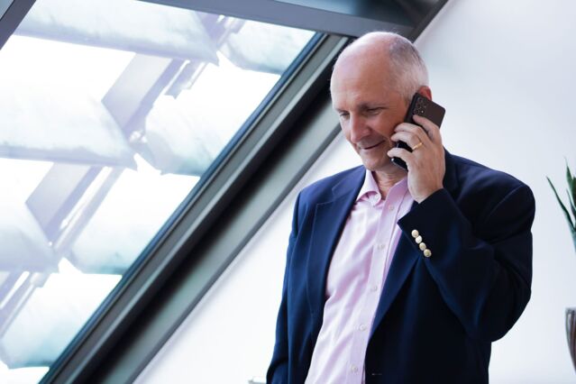 A man in a blue suit and pink shirt holds a smartphone to his ear and smiles. He has his gaze slightly downward. On the left, a large, slanted skylight.