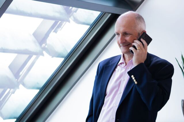 A man in a blue suit and pink shirt holds a smartphone to his ear and smiles. He has his gaze slightly downward. On the left, a large, slanted skylight.