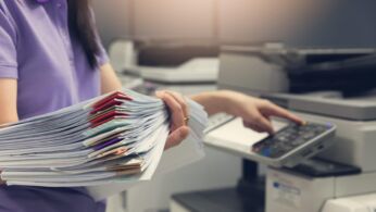 Bussinesswoman using copier machine to copy heap of paperwork in office.