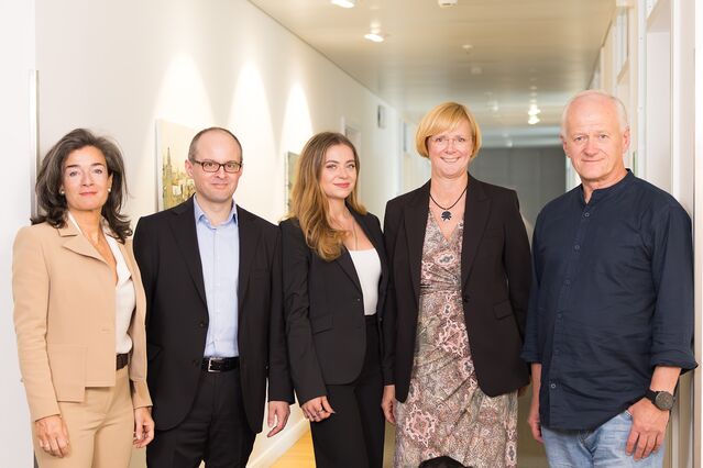 In this team photo in the office, two women are smiling from the left, a man, another woman and again a man. They have different attitudes and seem very competent.