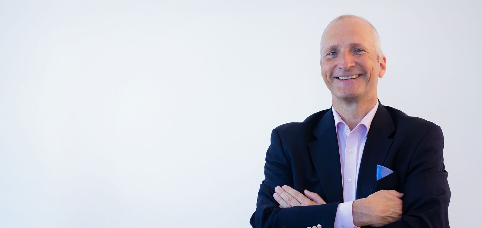 The photo shows a man in a blue suit with a pink shirt, smiling and crossing his arms in front of his chest.