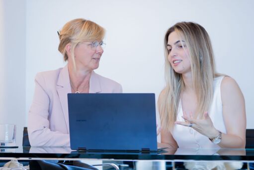 Zwei Damen sitzen vor einem Laptop, der auf einem einem Glasschreibtisch steht, und diskutieren.