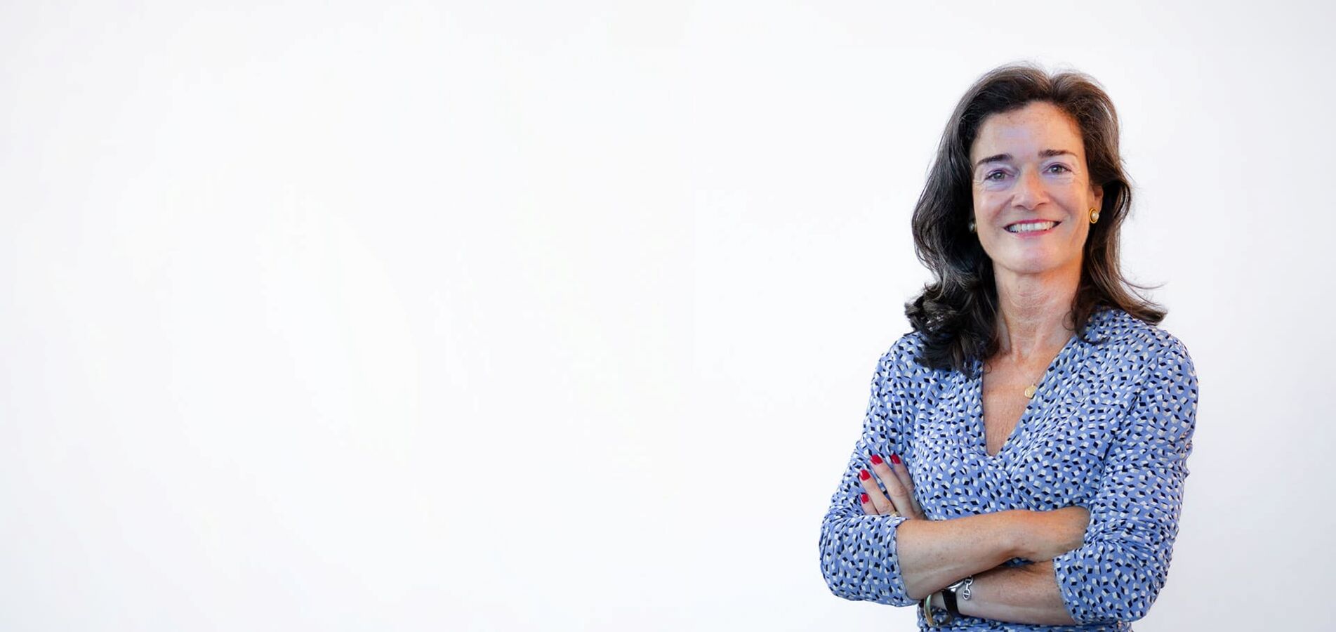 The photo shows an elderly lady with long dark brown hair wearing a white and blue dotted dress. She crosses her arms in front of her chest and smiles friendly into the camera.