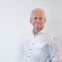 Ein Porträtfoto vom Geschäftsführer Arno Schlösser von DP-Dock. Er steht vor einer hellgrauen Wand und trägt ein weißes Hemd.