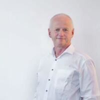 A portrait photo of the managing director Arno Schlösser of DP-Dock. He stands in front of a light gray wall and wears a white shirt.