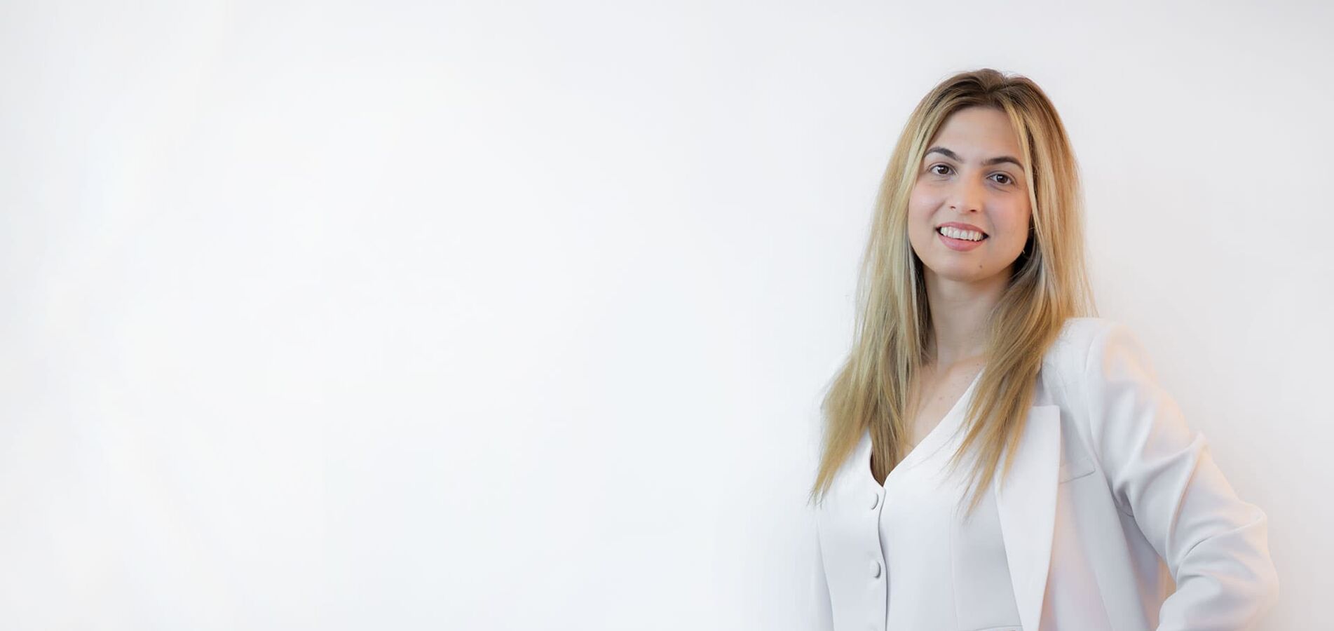 The photo shows a woman with long blond hair, wearing a white suit and smiling friendly into the camera.