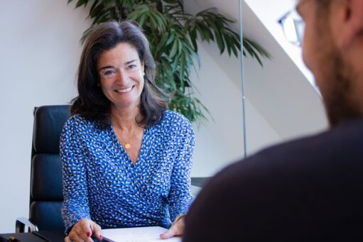 An older, friendly lady with dark hair wears a white dotted blouse and sits on an office chair at a desk, smiling. A man sits across from her. You can see him slightly from behind.