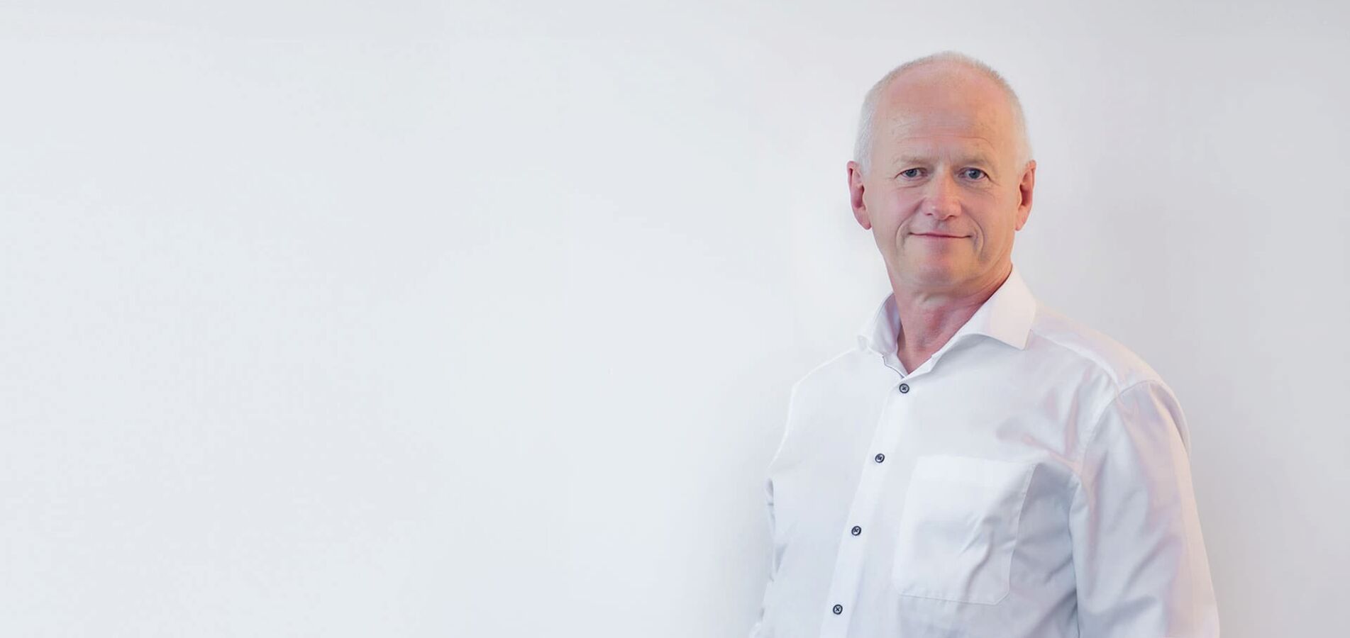 A portrait photo of the managing director Arno Schlösser of DP-Dock. He stands in front of a light gray wall and wears a white shirt.