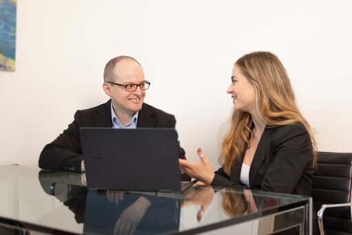 DP-Dock - Data Protection - Wir beraten Sie gerne! Zwei Damen sitzen vor einem Laptop, der auf einem einem Glasschreibtisch steht, und diskutieren.