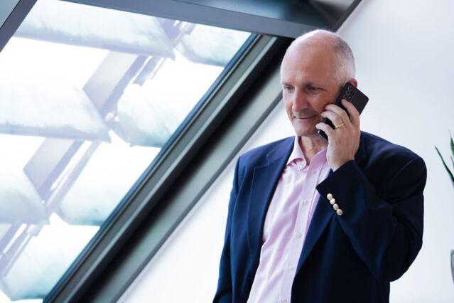 A man in a blue suit and pink shirt holds a smartphone to his ear and smiles. He has his gaze slightly downward. On the left, a large, slanted skylight.