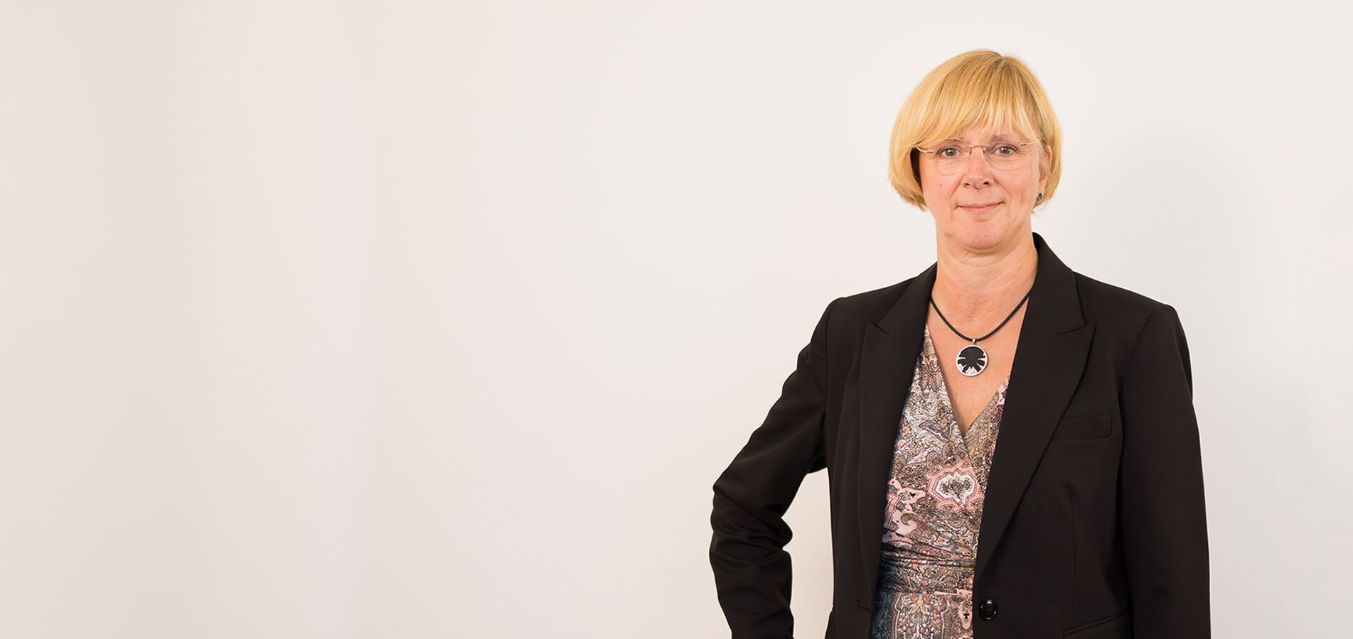 Ein Porträtfoto einer Dame im mittleren Alter vor einer hellgrauen Wand. Sie trägt eine Bluse und einen Blazer in rosé und verschränkt die Armen. Sie hat ihre blonden Haare hochgebunden, trägt eine Brille und lächelt in die Kamera.